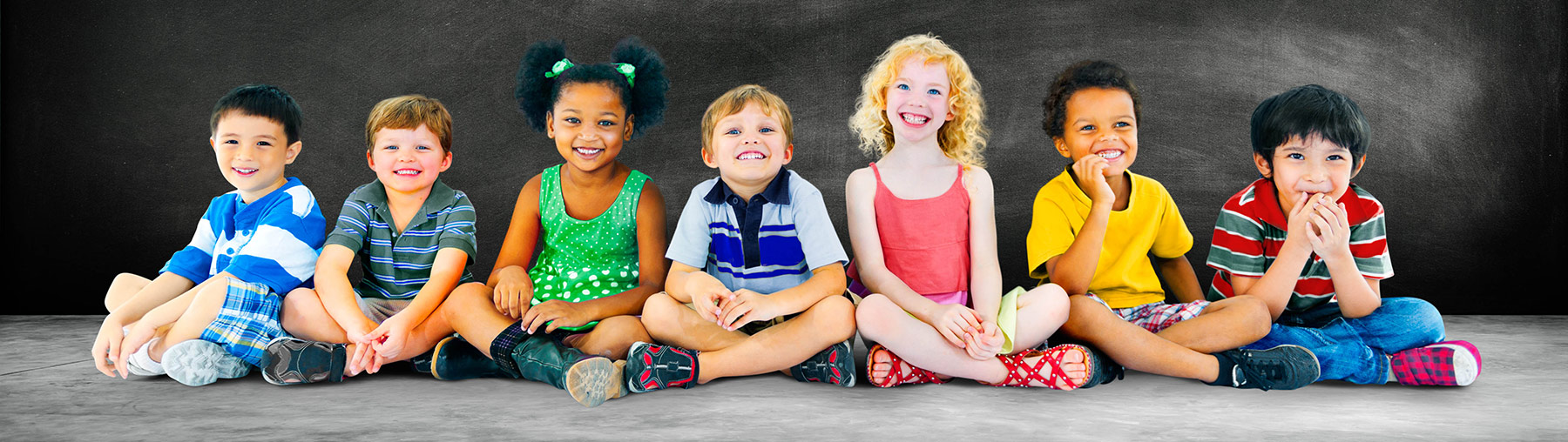 6 young children in colorful clothing, sitting on the ground in  front of a black board.